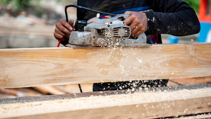 Male carpenter hand using an electric planer on wooden plank at construction site. Woodworker planing timber in his workshop with sawdust spreading around. Lumber industry and carpentry work tool
