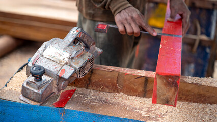 Rustic electric planer on workbench with carpenter hand squaring timber behind. Woodworker planing wooden plank at construction site. Carpentry work tool and lumber industry concept