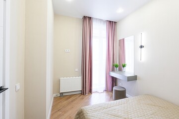 cozy room with beige walls, wooden floor, and pink curtains. A vanity table with a mirror and a stool sits by the window. A bed with a quilted cover is in the foreground