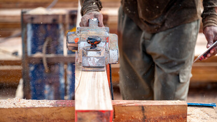 Male carpenter hand using an electric planer on wooden plank in workshop. Woodworker planing timber at construction site with sawdust spreading around. Work tool for carpentry. Lumber industry concept