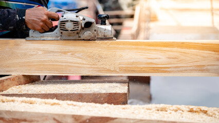 Male carpenter hand using an electric planer on wooden plank in workshop. Woodworker planing timber at construction site with sawdust spreading around. Work tool for carpentry. Lumber industry concept