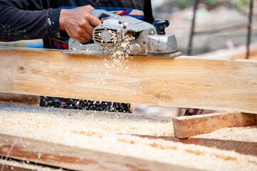 Male carpenter hand using an electric planer on wooden plank in workshop. Woodworker planing timber at construction site with sawdust spreading around. Work tool for carpentry. Lumber industry concept