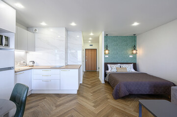 bright studio apartment featuring a white kitchen, herringbone floors, and a cozy bedroom area with green patterned wallpaper