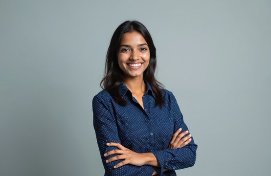 Smiling young Indian woman posing in studio. Confident businesswoman or student with crossed arms looks at camera. Successful, happy, cheerful woman in formal shirt on grey background.
