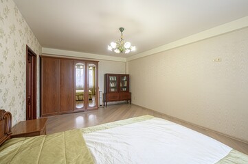 Bedroom with wood floors, floral wallpaper, large wardrobe, and a bed with a white and green cover. Chandelier on the ceiling