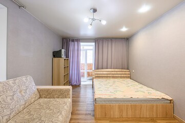 Cozy room with a patterned sofa, wooden bed, and light purple curtains. Simple decor, bright lighting.