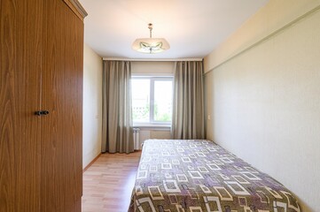 Obraz premium small, cozy bedroom with a single bed, wooden wardrobe, window with curtains, and a simple ceiling light