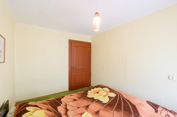 room with pale yellow walls, a brown door, and a bed with a floral blanket. A light fixture hangs...
