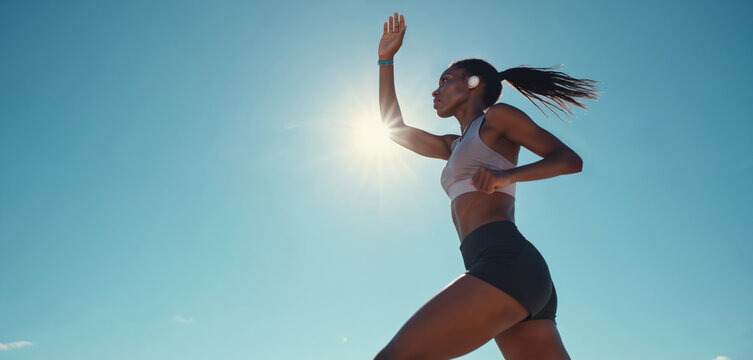 Young black woman runs outdoors on blue sky background wearing earbuds. Runner in sports clothing, fitness, health, marathon or cardio. Active lifestyle, exercise, wellness concept. - Powered by Adobe