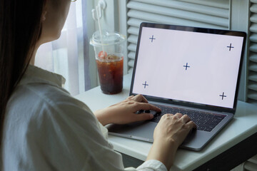 laptop blank white screen mockup. woman using hand typing on keyboard laptop at home for work or entertainment on day time with ice coffee on the table near window with light filtering curtains