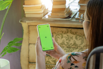 cell phone blank green screen mockup. woman holding mobile phone in hand, using smartphone at living room for work contact or entertainment. room decorate with vintage cabinet, book, Monstera tree