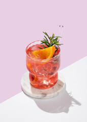 Red orange cocktail  with ice and rosemary on a marble stand on a  pink white background