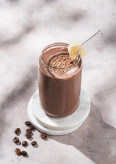 Chocolate coffee smoothie with banana in a glass on a light  background with morning shadow.