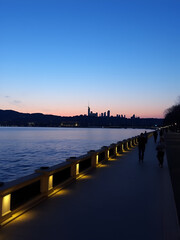 Fototapeta premium Evening stroll along illuminated lakeside promenade, city skyline reflecting in calm water, twilight sky.