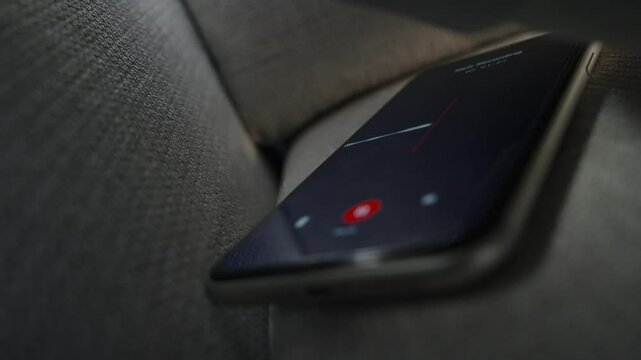 Smart phone with voice recording application is covertly grabbing conversation. Mobile phone hidden under and between couch or sofa pillows or bed. Wiretapping app or surveillance evidence gathering