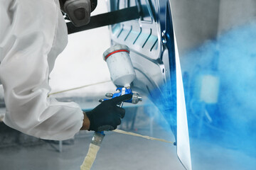 Professional male car painter sprays paints car part to blue color in garage by airbrush. Man works...