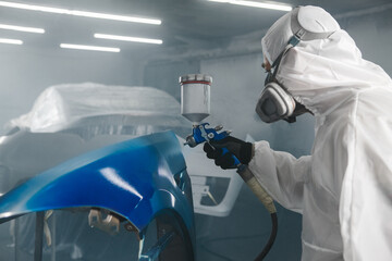 Worker in protective mask and jumpsuit overall spraying base coat of paint on car body elements in...