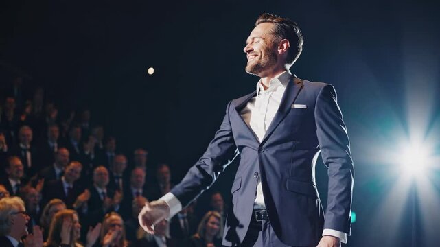 Smiling businessman standing on stage, gesturing to audience applauding his excellent presentation, bright spotlight shining on him in large auditorium