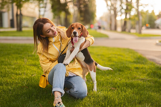 A joyful scene of a young woman interacting with her beagle dog on a grassy area, enjoying the outdoors under the warm sunlight.