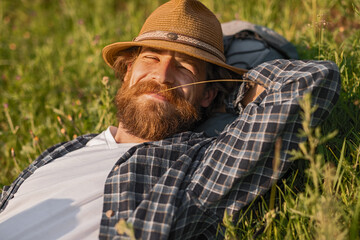 A bearded man in a hat relaxes in a grassy field, enjoying the serene outdoors and sunlight. His...