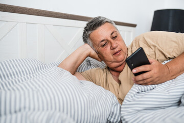 Happy mature grey hair woman with smartphone laying in a bed in the morning.
