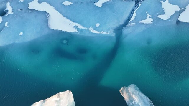 Aerial drone shot of large icebergs floating in deep blue glacial lagoon in Iceland. Clear water reveals submerged ice. Frozen textures and light reflections create peaceful winter scene from above