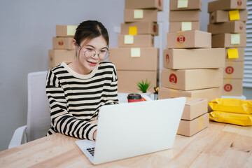 A young woman manages an online business, holding packages and working on a laptop surrounded by stacked boxes.