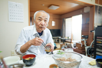 In Japan, a Japanese man in his late 70s wears a white shirt and sits in a modest apartment room, quietly eating breakfast. Morning light gently fills the space, starting the day in peace.