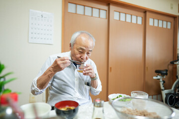 In Japan, a Japanese man in his late 70s wears a white shirt and sits in a modest apartment room, quietly eating breakfast. Morning light gently fills the space, starting the day in peace.