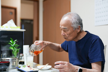 In a small Japanese apartment, a Japanese man in his late 70s relaxes in T-shirt pajamas. He sits alone in the evening, sipping whiskey and enjoying a calm, quiet moment after a long day of solitude.