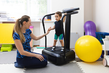 Little boy during vibration therapy standing on vibration platform with her assistant in a rehabilitation center