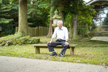 Fototapeta premium In rural Japan, a Japanese man in his late 70s takes a quiet rest at a countryside shrine surrounded by lush nature. Wearing a white shirt and hat, he sits calmly, enjoying the early summer air.