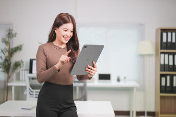 Happy Asian businesswoman standing in a modern office, using a digital tablet while pointing at the screen, smiling confidently and engaging in her work