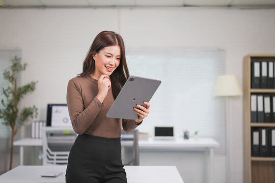 Young Asian businesswoman smiling while using a digital tablet in a modern office, contemplating her job and strategizing for future success in her career