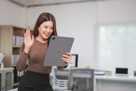 Young happy asian businesswoman waving at tablet during a video conference call in her office, she is using a modern device for online communication and remote work - Powered by Adobe
