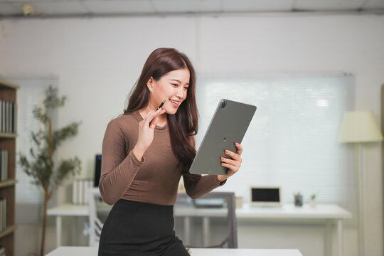 Young businesswoman engaging in a video call while sitting at her modern office desk, holding a pen and smiling, radiating confidence and professionalism in her stylish workspace