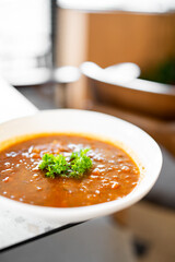A close-up of bowl of hearty tomato soup garnished with fresh parsley. The rich, vibrant red color of the soup highlights its fresh ingredients. The dish is presented on a table in a well-lit setting