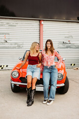 Two young women leaning on red convertible car enjoying road trip