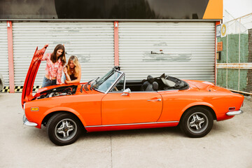 Two young women checking the engine of a vintage red cabriolet car