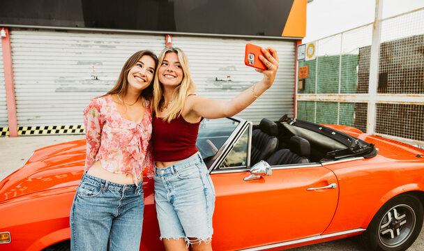 Two young women taking a selfie with a red convertible car during road trip