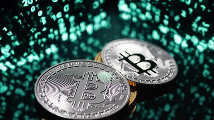Two shiny silver and gold bitcoin cryptocurrency coins rest on a reflective black surface with glowing green computer code numbers floating in the background - Powered by Adobe