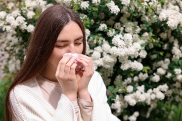 Woman with paper tissue suffering from seasonal pollen allergy near blossoming shrub on spring day,...