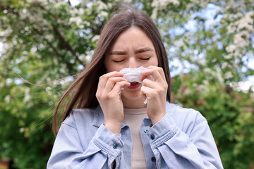 Woman with paper tissue suffering from seasonal pollen allergy near blossoming tree on spring day