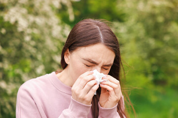 Woman with paper tissue suffering from seasonal pollen allergy near blossoming shrub on spring day