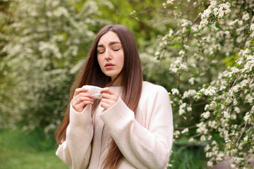 Woman with paper tissue suffering from seasonal pollen allergy near blossoming shrub on spring day