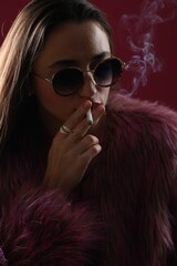 Beautiful woman in fur coat smoking cigarette on dark pink background, closeup