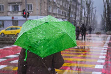 Person with green umbrella walking on city crosswalk during snowfall, urban winter scene. Bright...