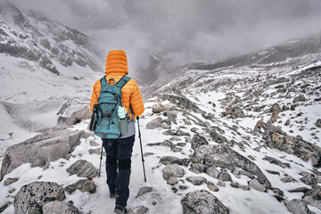 Hiker with backpack walks along snowy trail through mountain pass against backdrop of an approaching snow storm in Himalayas.