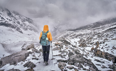 Hiker with backpack walks along snowy trail through mountain pass against backdrop of dramatic mountains and clouds.