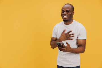 Young african american man laughing and holding smartphone on yellow background